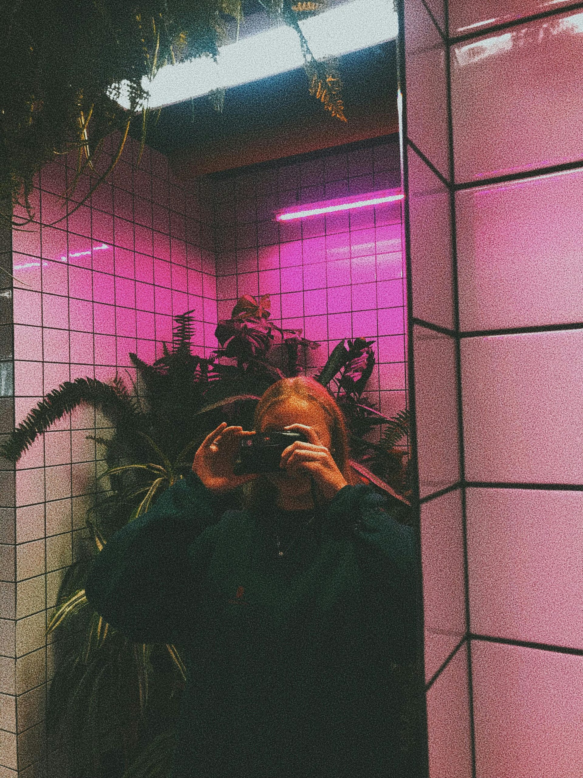 Young female taking selfie on photo camera under pink neon light in mirror